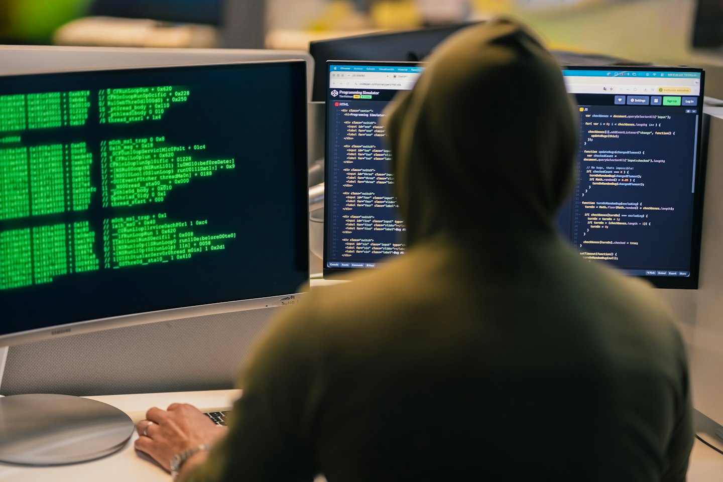 Programmer wearing a hoodie works on two monitors displaying code.